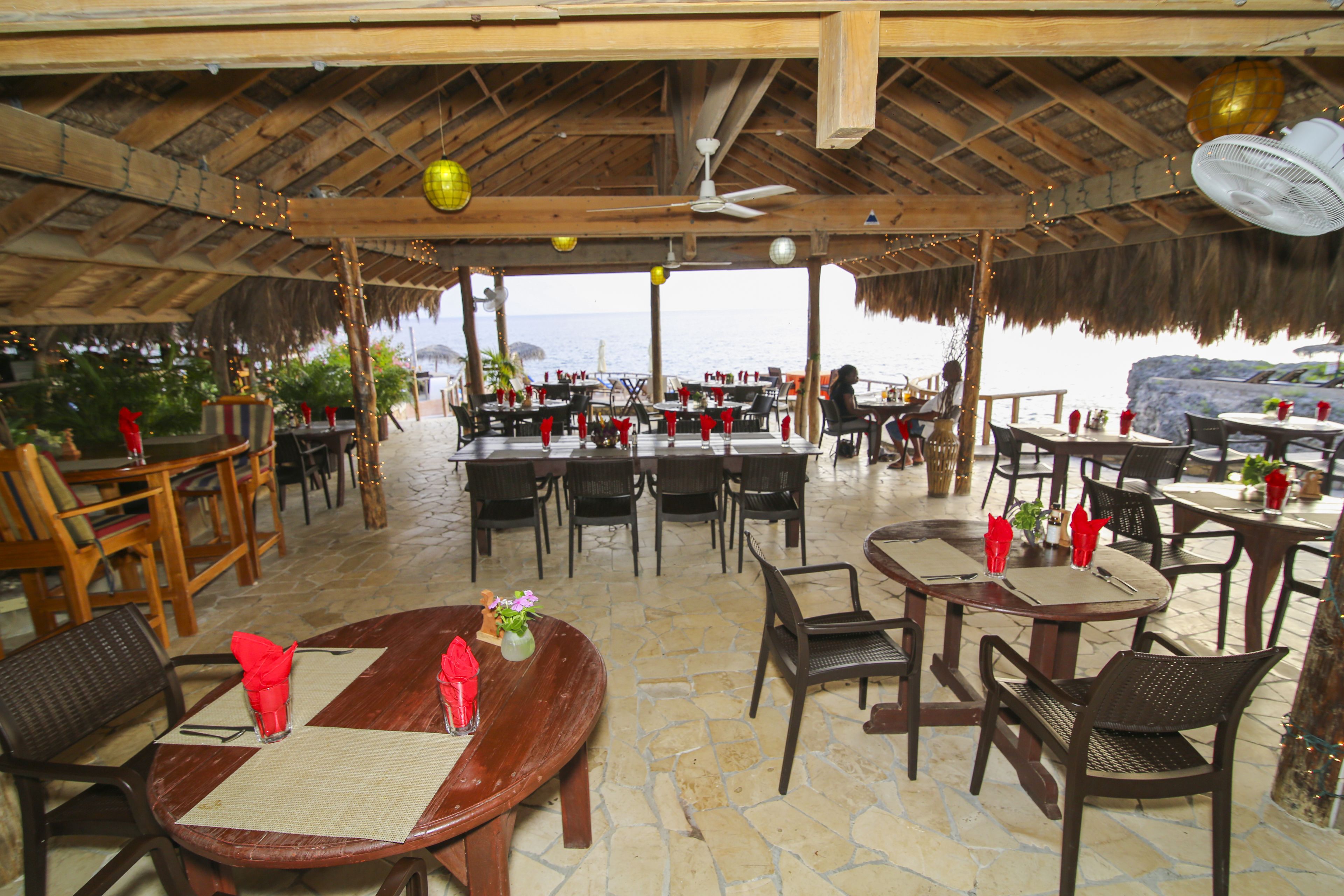 An outdoor restaurant with wooden tables and chairs, a thatched roof, and a view of the sea.