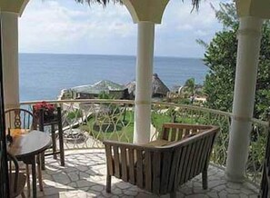 A covered outdoor seating area with a view of the ocean and a thatched-roof building in the background.