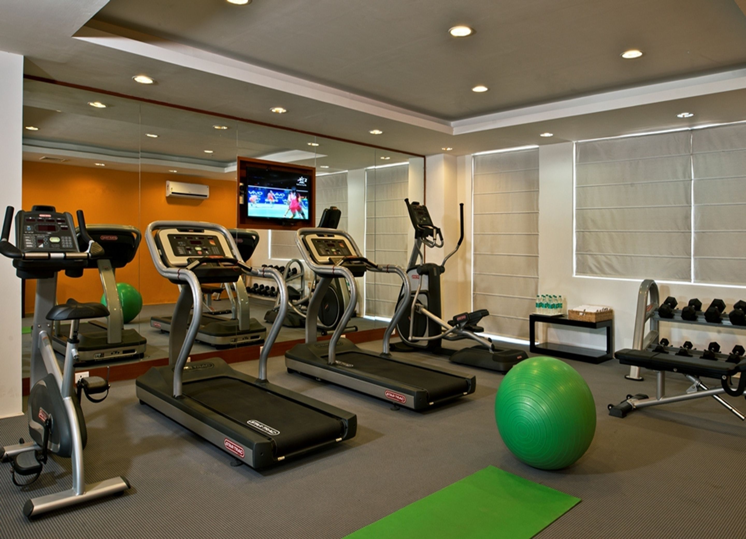 A gym with treadmills, elliptical machines, a green exercise ball, and a TV screen displaying a sports event.