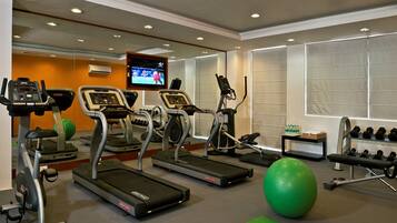 A gym with treadmills, elliptical machines, a green exercise ball, and a TV screen displaying a sports event.