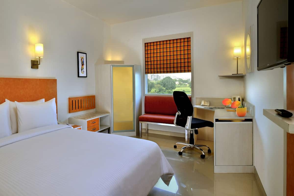 A hotel room with a large bed, a desk with a chair, a television, and a window with a view of greenery.