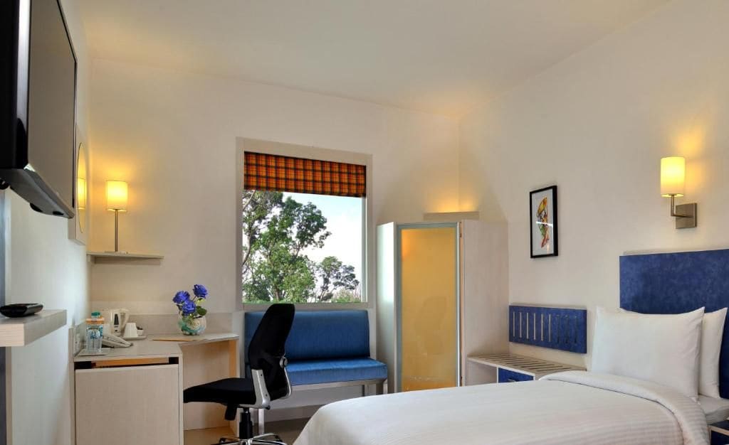 Una habitación de hotel con cama, escritorio, silla, armario y ventana con vista a árboles.