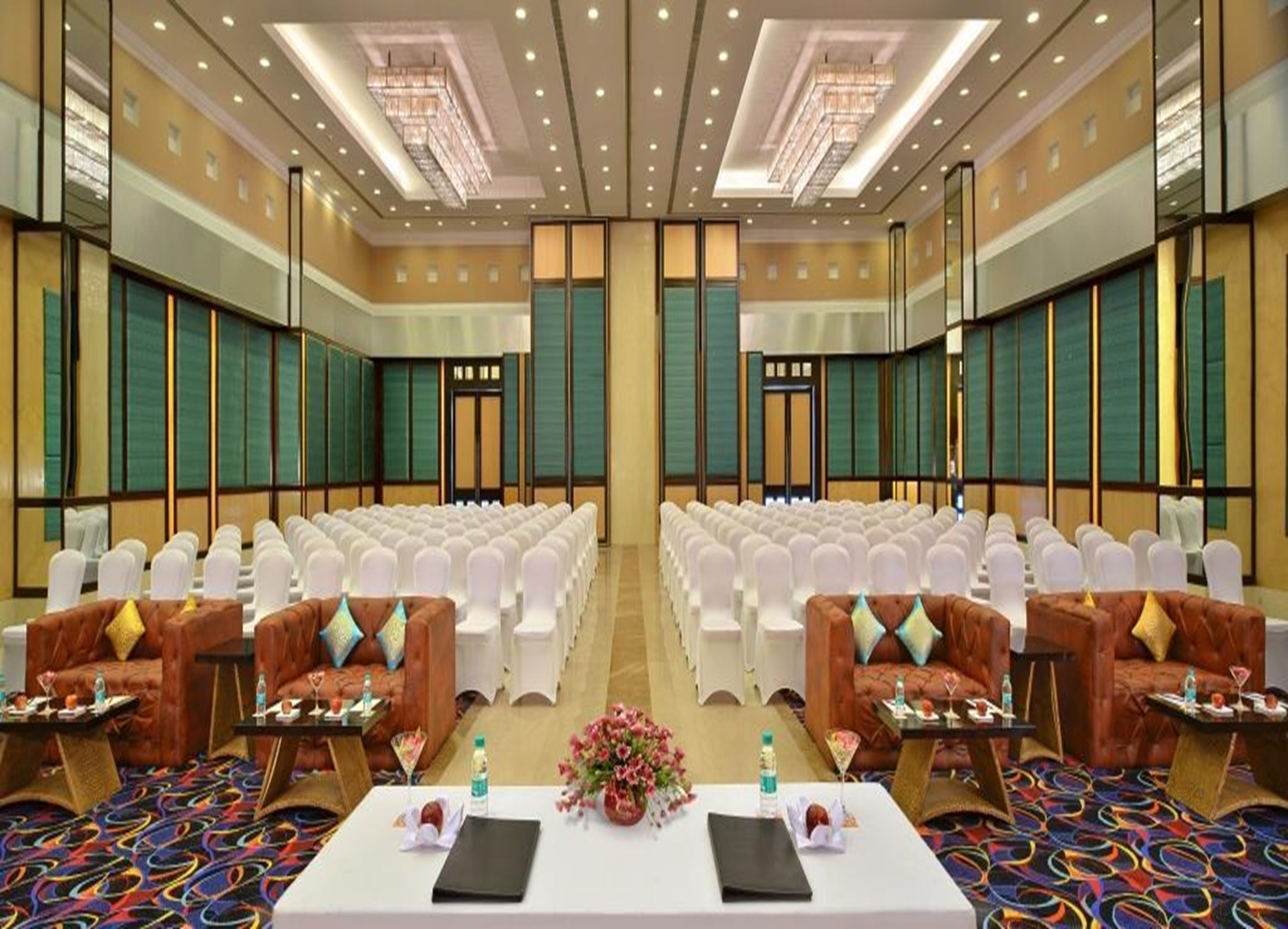 A conference room with rows of white chairs, a central table with a floral arrangement, and two sofas with side tables.