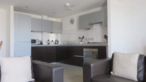 A modern kitchen with grey cabinets, a microwave, kettle, and a clock on the wall. There are two grey sofas in the living area.