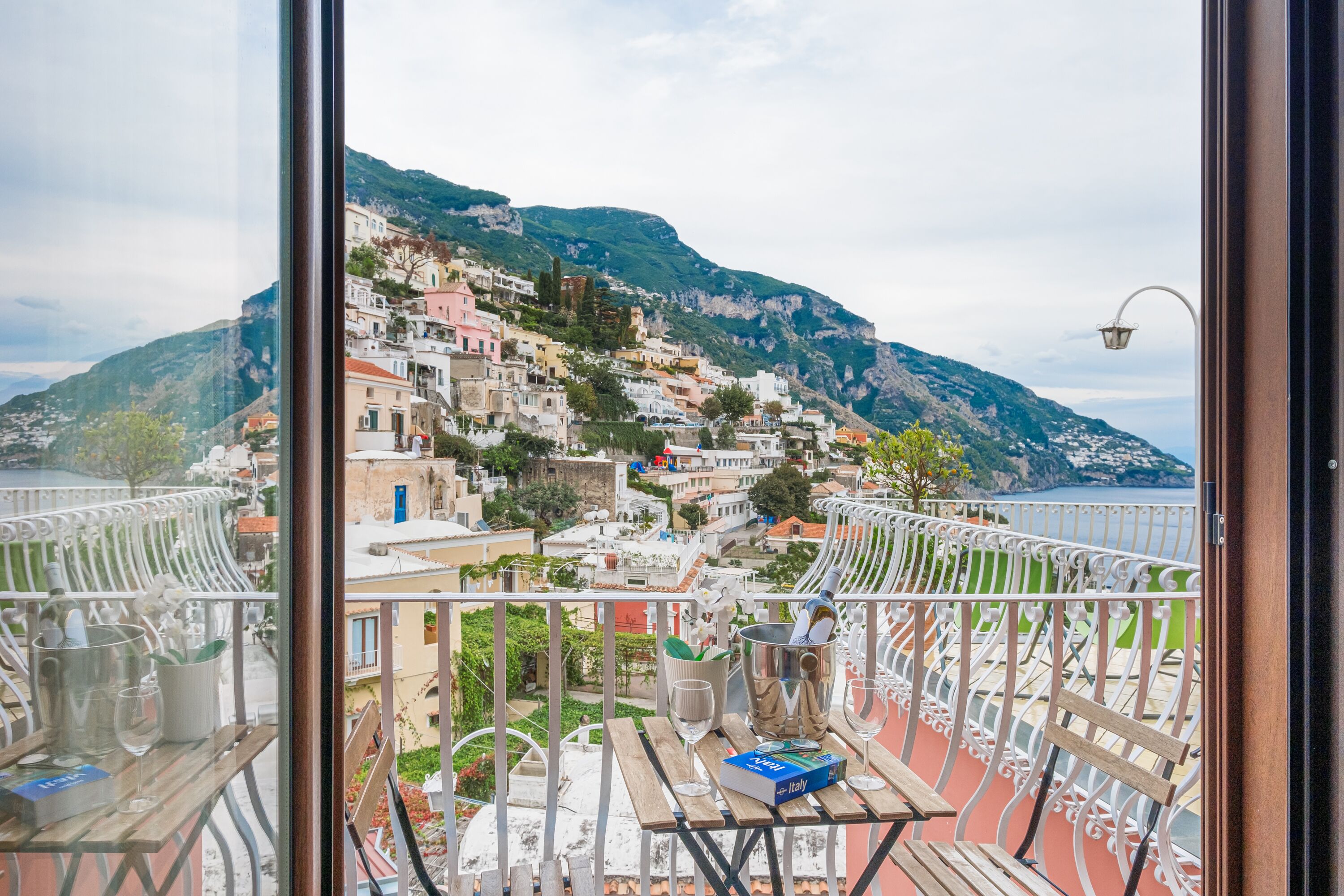 A balcony with a view of a hillside town, a table with a book and glasses, and a metal bucket.