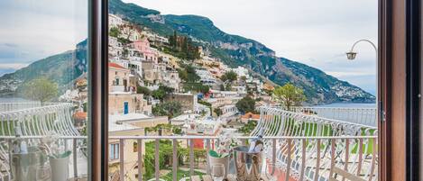 A balcony with a view of a hillside town, a table with a book and glasses, and a metal bucket.