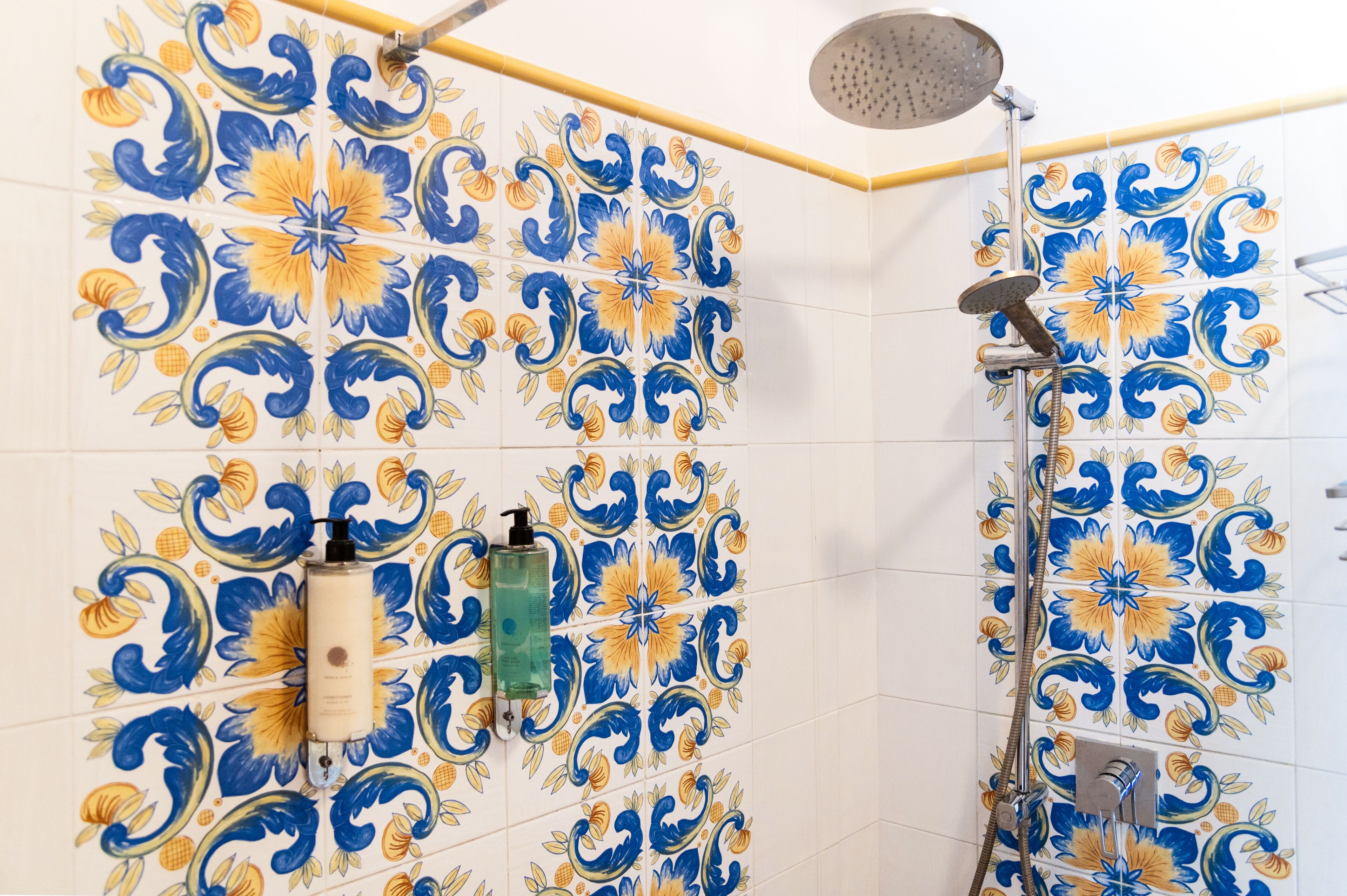 Un baño con baldosas decorativas que presentan motivos florales en azul y amarillo, un cabezal de ducha manual y un dispensador de jabón montado en la pared.