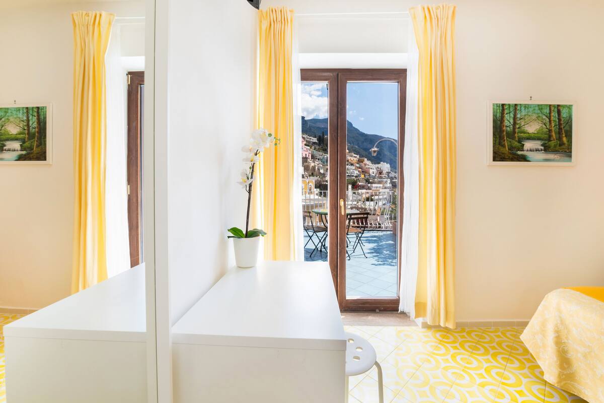 Una habitación con un ventanal grande, un escritorio blanco, una planta en maceta y una cama con colcha de cuadros amarillos y blancos.