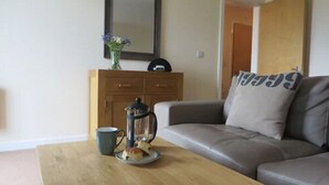A living room with a sofa, a coffee table with a cup and a teapot, and a cabinet with a vase of flowers. - Citystay Living - Warren Close (Cambridge)