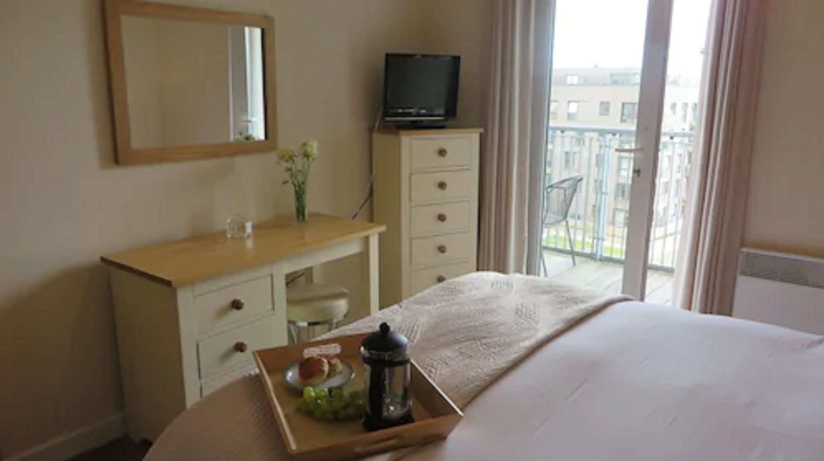 Hotelkamer met een bed, een kaptafel, een televisie op een standaard, een raam met uitzicht op gebouwen, en een dienblad met fruit en een lantaarn.