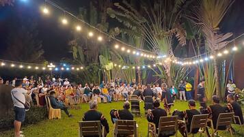 Reunión al aire libre con gente sentada en sillas, rodeada de luces colgantes y palmeras.