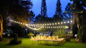 Un espacio para eventos al aire libre con mesas, sillas, luces colgantes y un vehículo al fondo.
