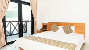 Habitación de hotel con una cama grande, cabecera de madera, dos almohadones estampados, una mesita de noche con un teléfono y una libreta, y vista a través de un ventanal corredero.