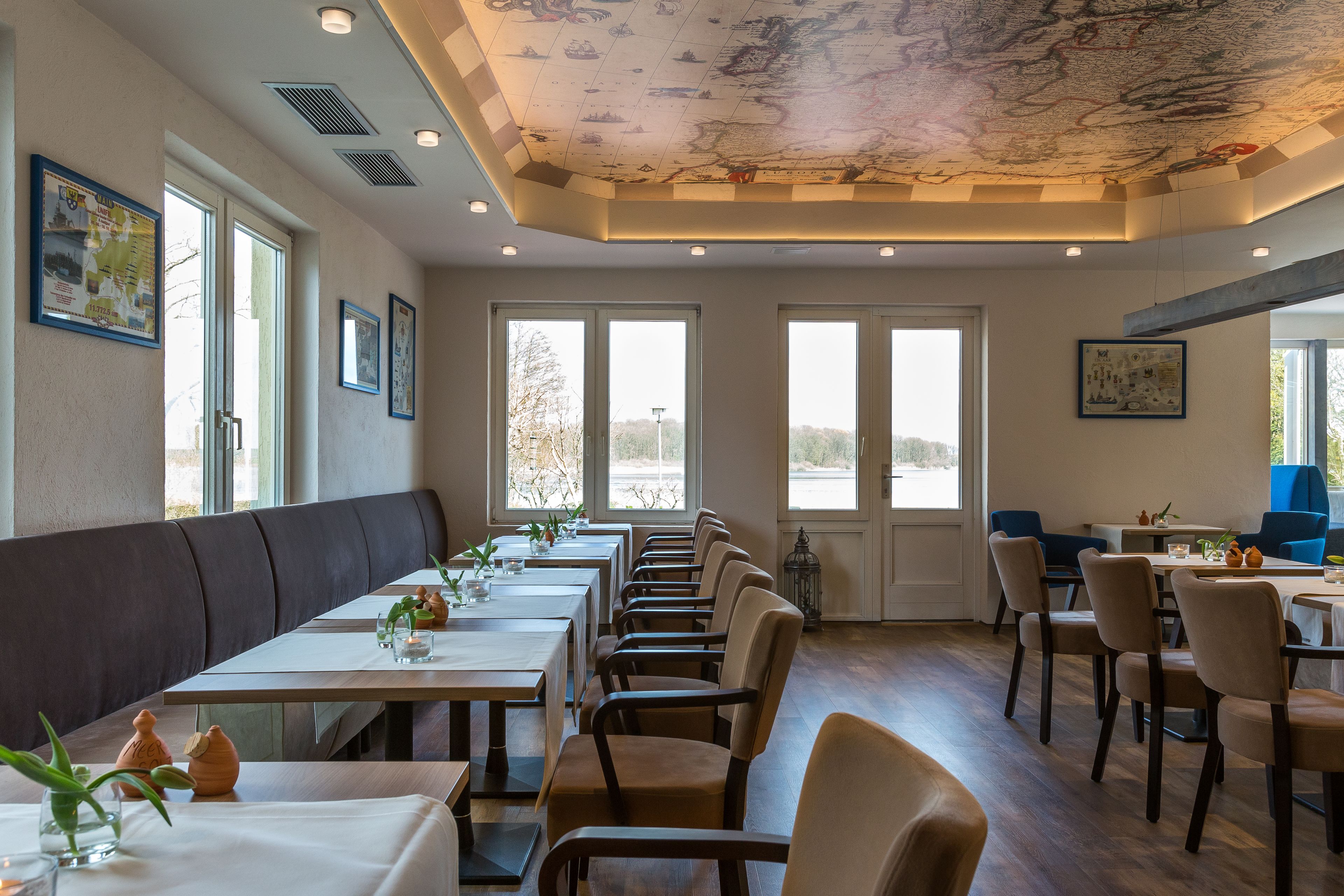 A restaurant with long tables and booths, large windows, and a ceiling with a map design.