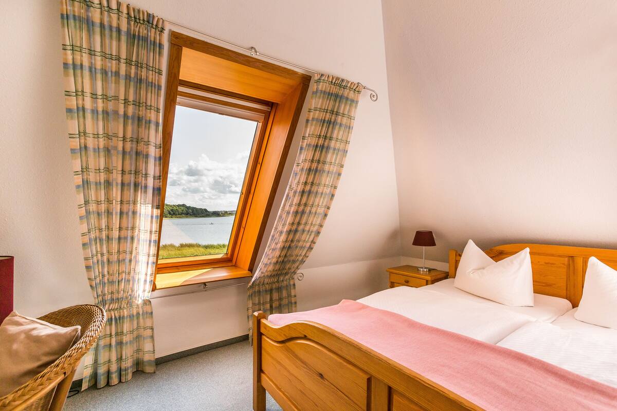 A bedroom with a wooden bed, a window with curtains, a chair, and a nightstand with a lamp.