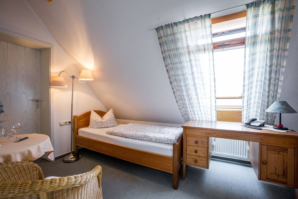A small, cozy room with a single bed, a desk with a lamp, a chair, and a window with curtains.