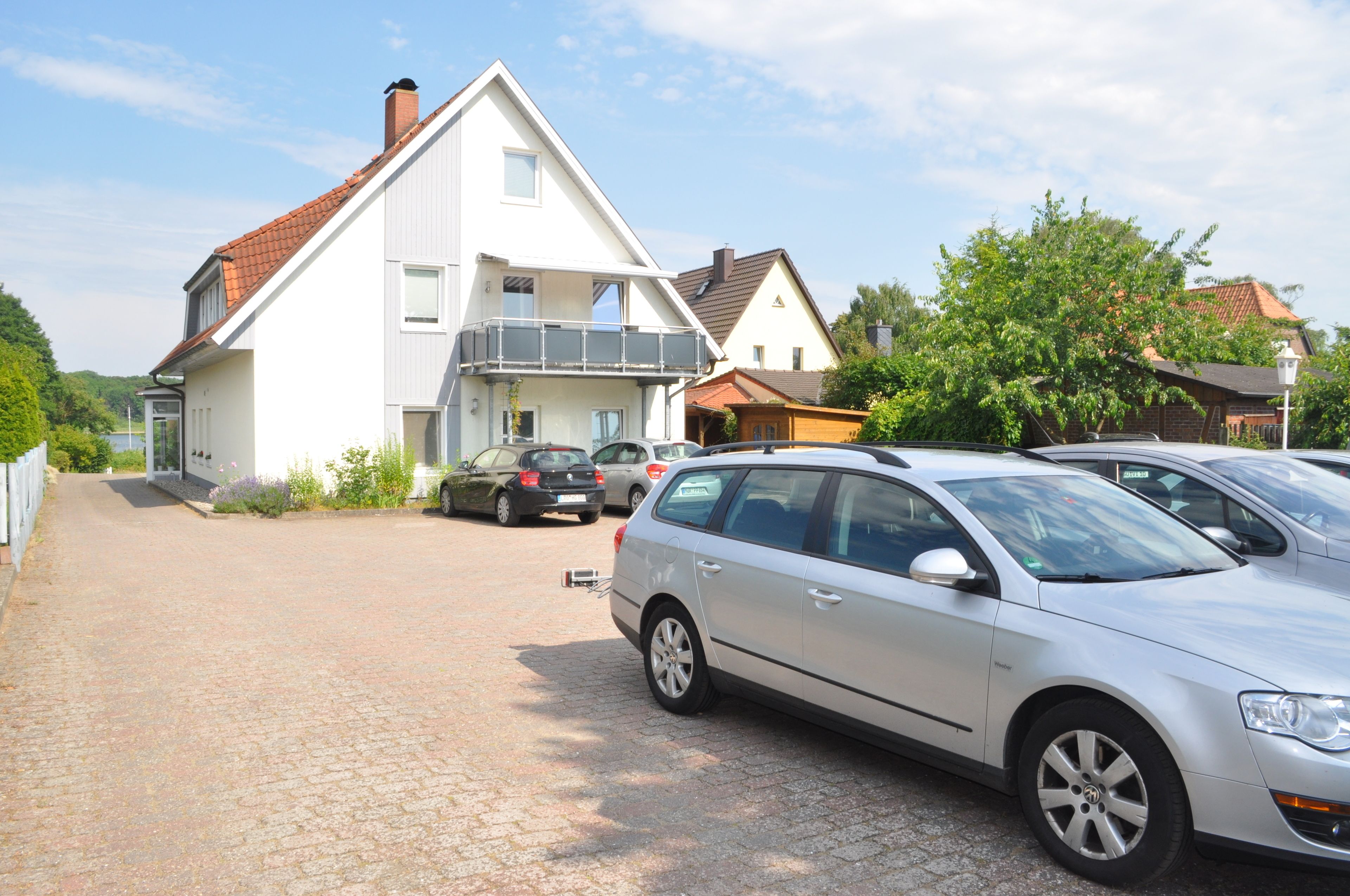 Rua residencial com uma casa branca com varanda, carros estacionados e entrada de automóveis com pedras.