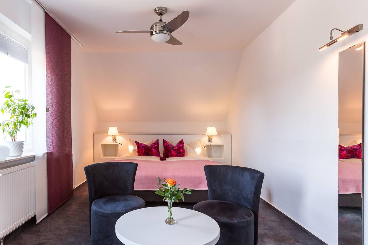 A hotel room with a bed, two bedside tables with lamps, a ceiling fan, a round table with a vase of flowers, and a potted plant by the window.