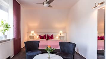 Hotellihuoneessa on sänky, kaksi yöpöytää, joissa on lamput, kattotuuletin, pyöreä pöytä, jossa on maljakko täynnä kukkia, ja ruukkukasvi ikkunan lähellä.