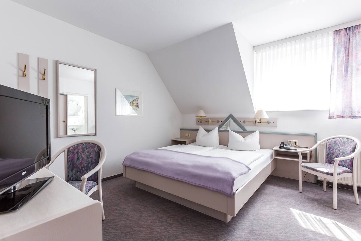 A modern hotel room with a bed, a desk with a laptop, a TV, and two chairs.