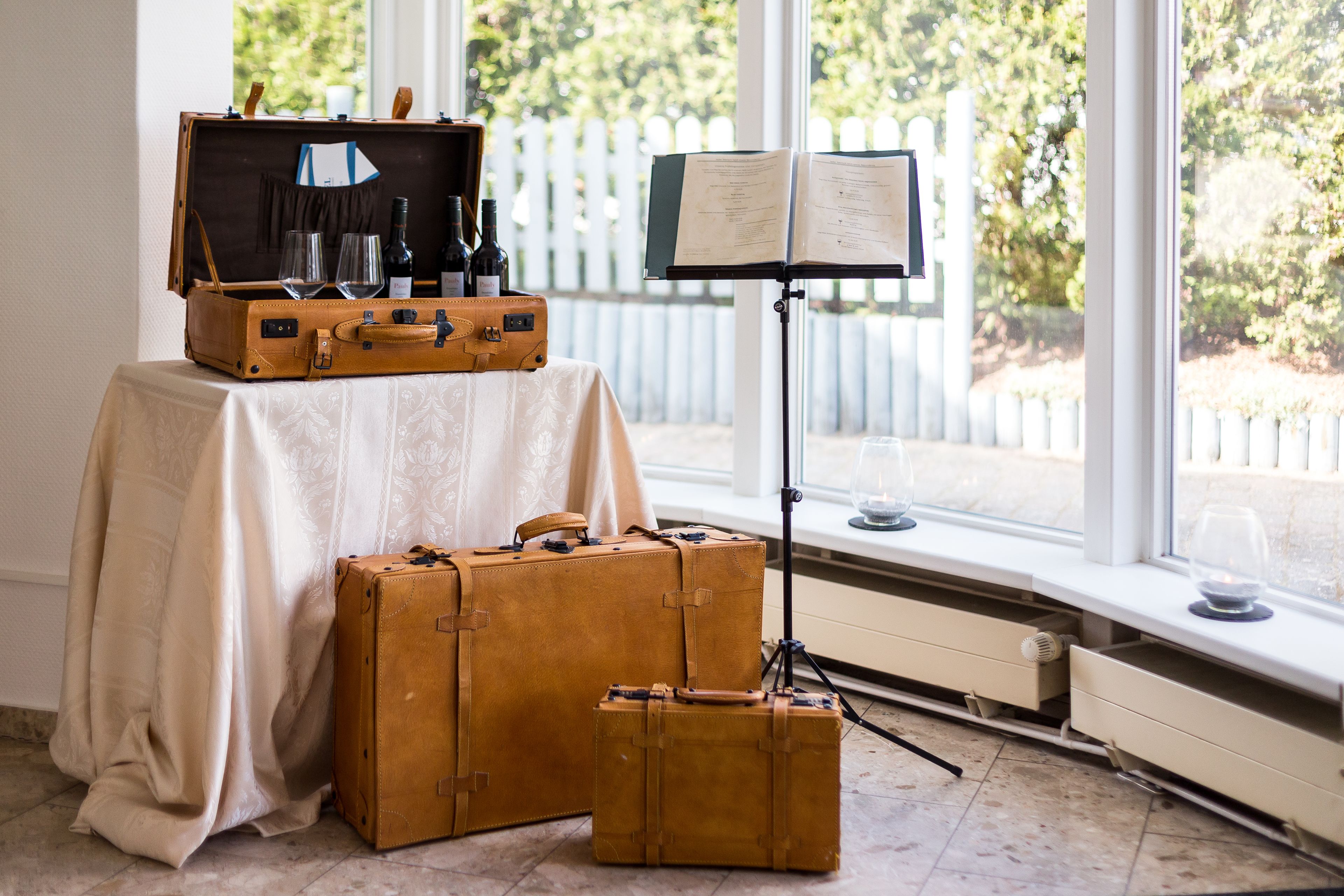 A room with a table covered by a white tablecloth, two brown suitcases, a music stand with sheet music, and a window with a view of greenery.