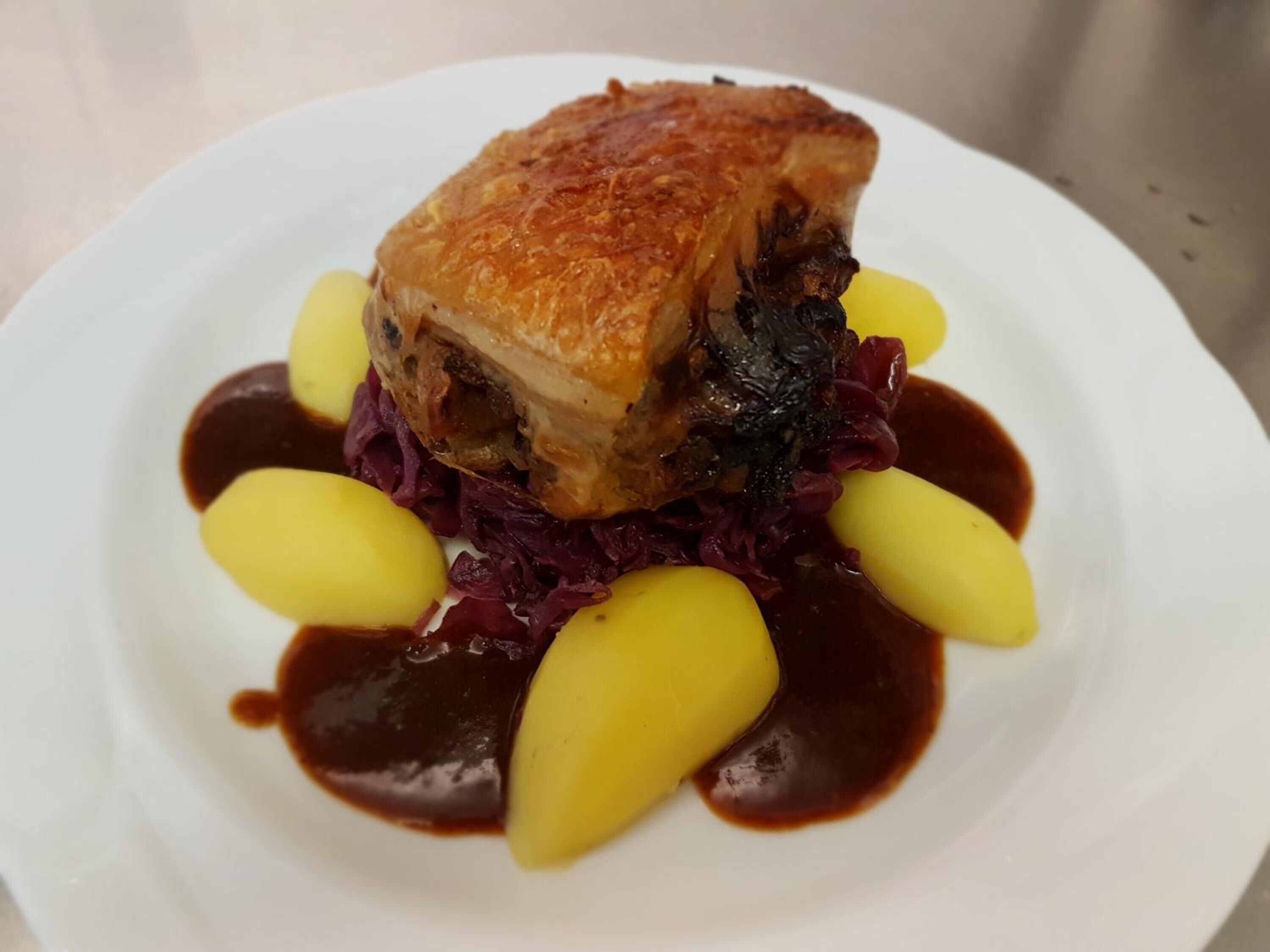Un plato con un codillo de cerdo asado, patatas amarillas y col roja, acompañado de salsa oscura.