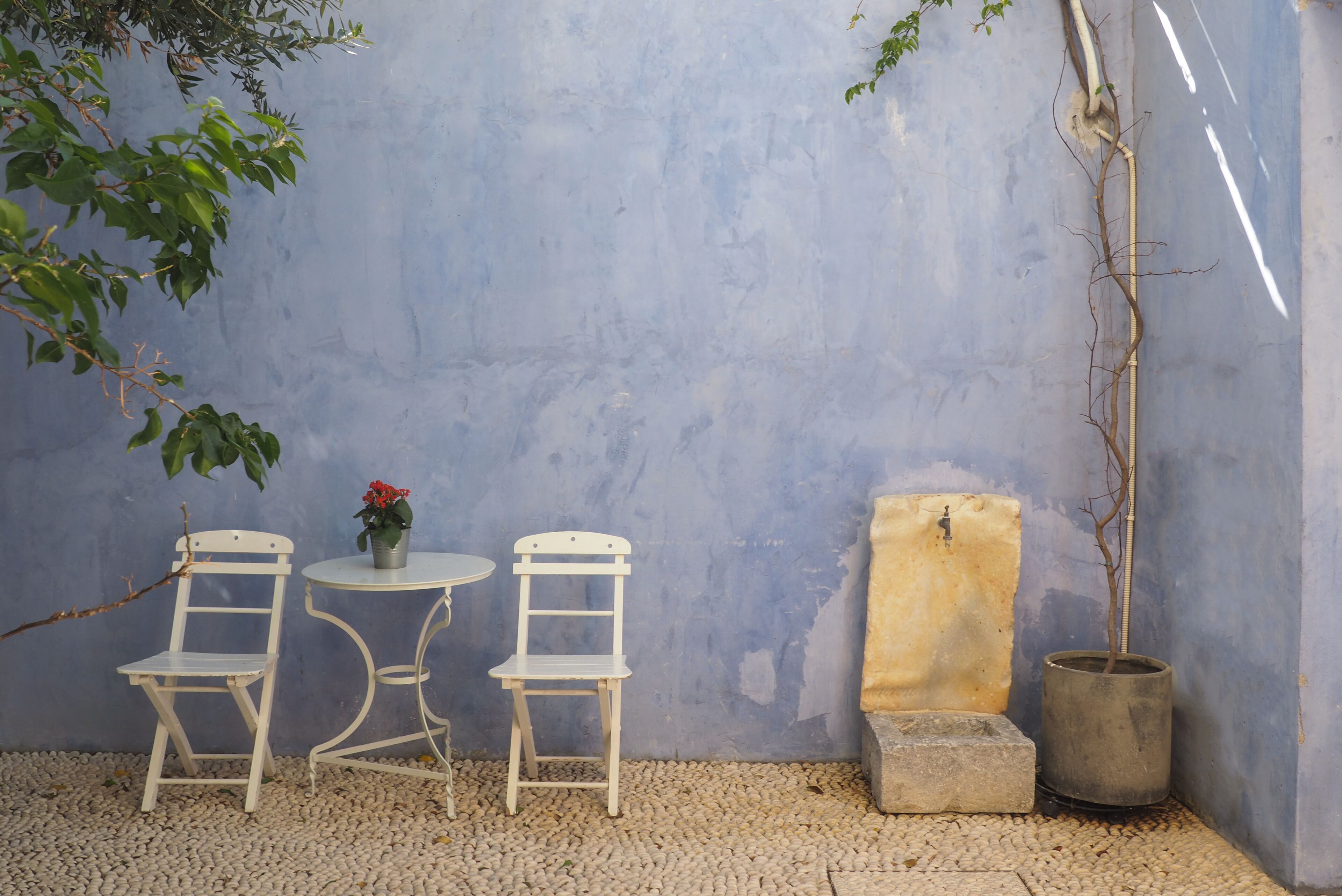 A small outdoor seating area with two white chairs and a round table, surrounded by pebbles and a blue wall.