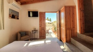 A bedroom with a bed, a TV mounted on the wall, a small table with a lamp, and a door leading to an outdoor patio area. - Saint George Studios (Rhodes)