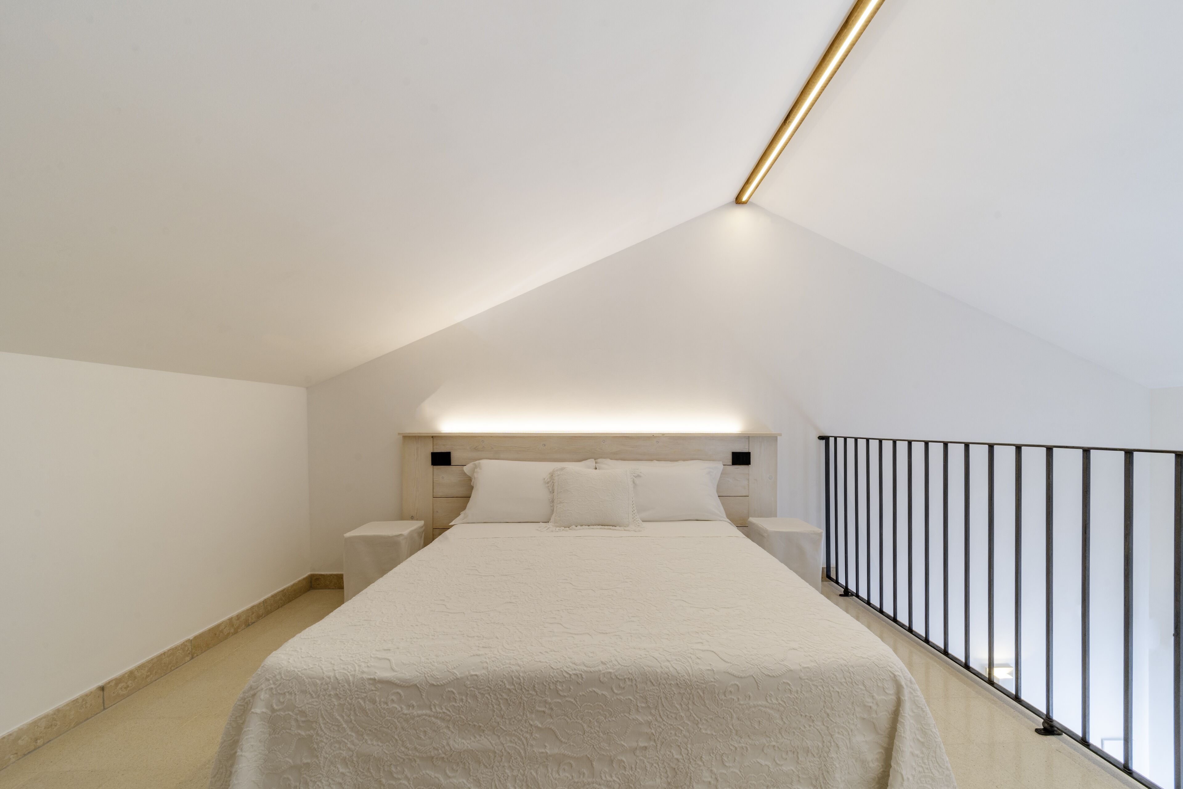 A bedroom with a sloped ceiling, a bed with a white bedspread, bedside tables, and a railing along the window.