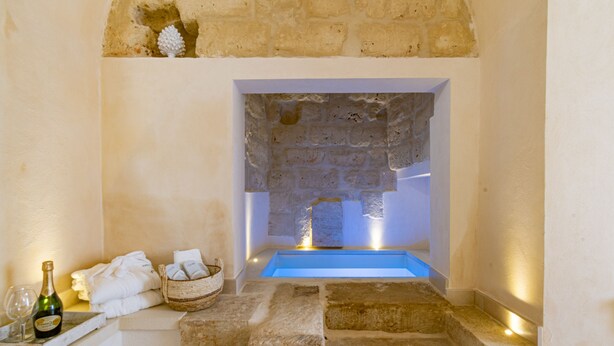 A stone-walled bathroom with a blue bathtub, a bottle of champagne, and towels on a shelf. - Masseria dei Monaci (Otranto)