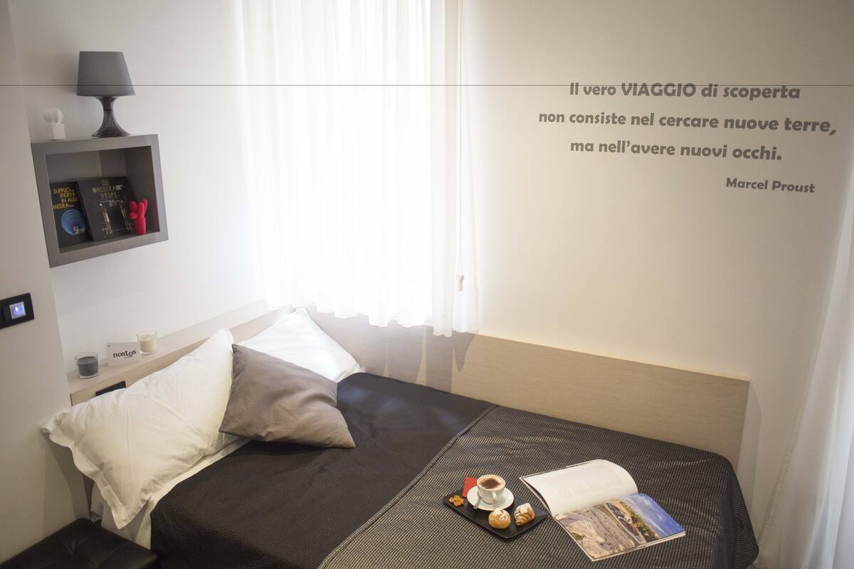 A hotel room with a bed, a bedside table with a lamp, a shelf with books and a red object, and a wall quote.