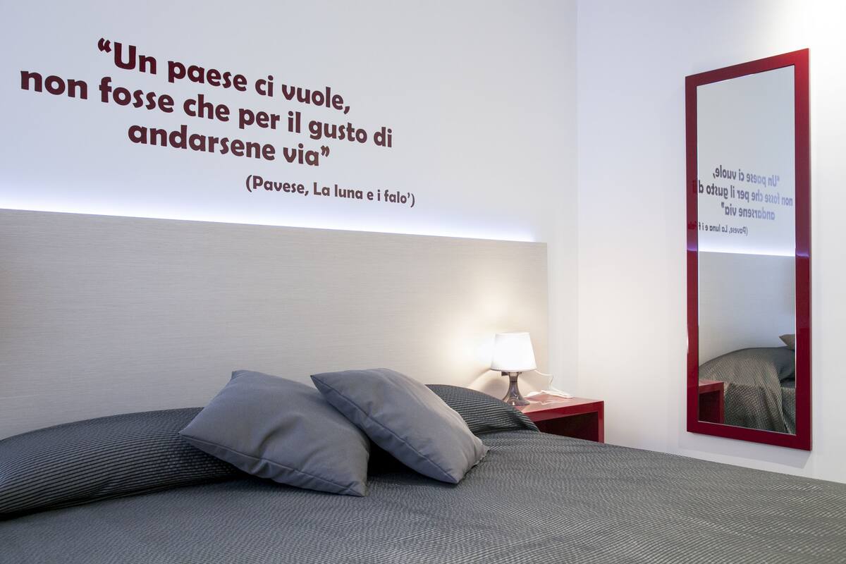 A modern hotel room with a large bed, a bedside table with a lamp, a wall-mounted mirror, and a quote in Italian.