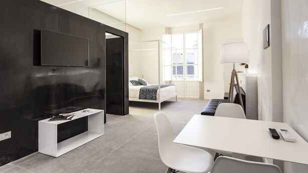 A modern hotel room with a flat-screen TV mounted on a dark wall, a white desk with a remote control, a bed with a black and white patterned bedspread, and a window with curtains. - Nostos rooms & Apartments (Syracuse)