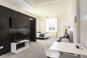 Una habitación de hotel moderna con una televisión de pantalla plana montada en una pared oscura, un escritorio blanco con un control remoto, una cama con una colcha a cuadros blanco y negro, y una ventana con cortinas.