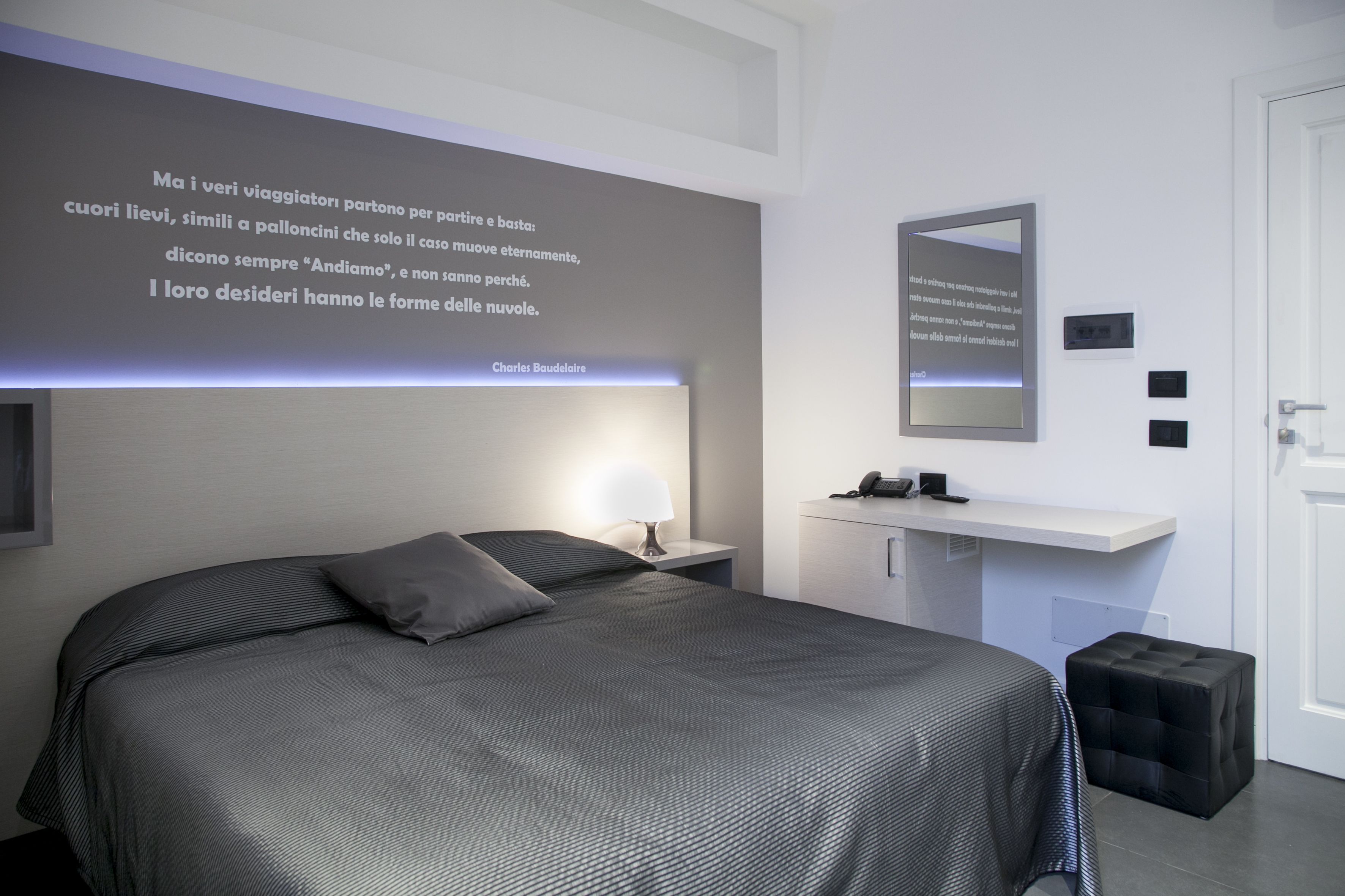 Un dormitorio moderno con una cama grande, una mesita de noche, un pequeño taburete, un aparador blanco, un espejo y una pared con una cita.