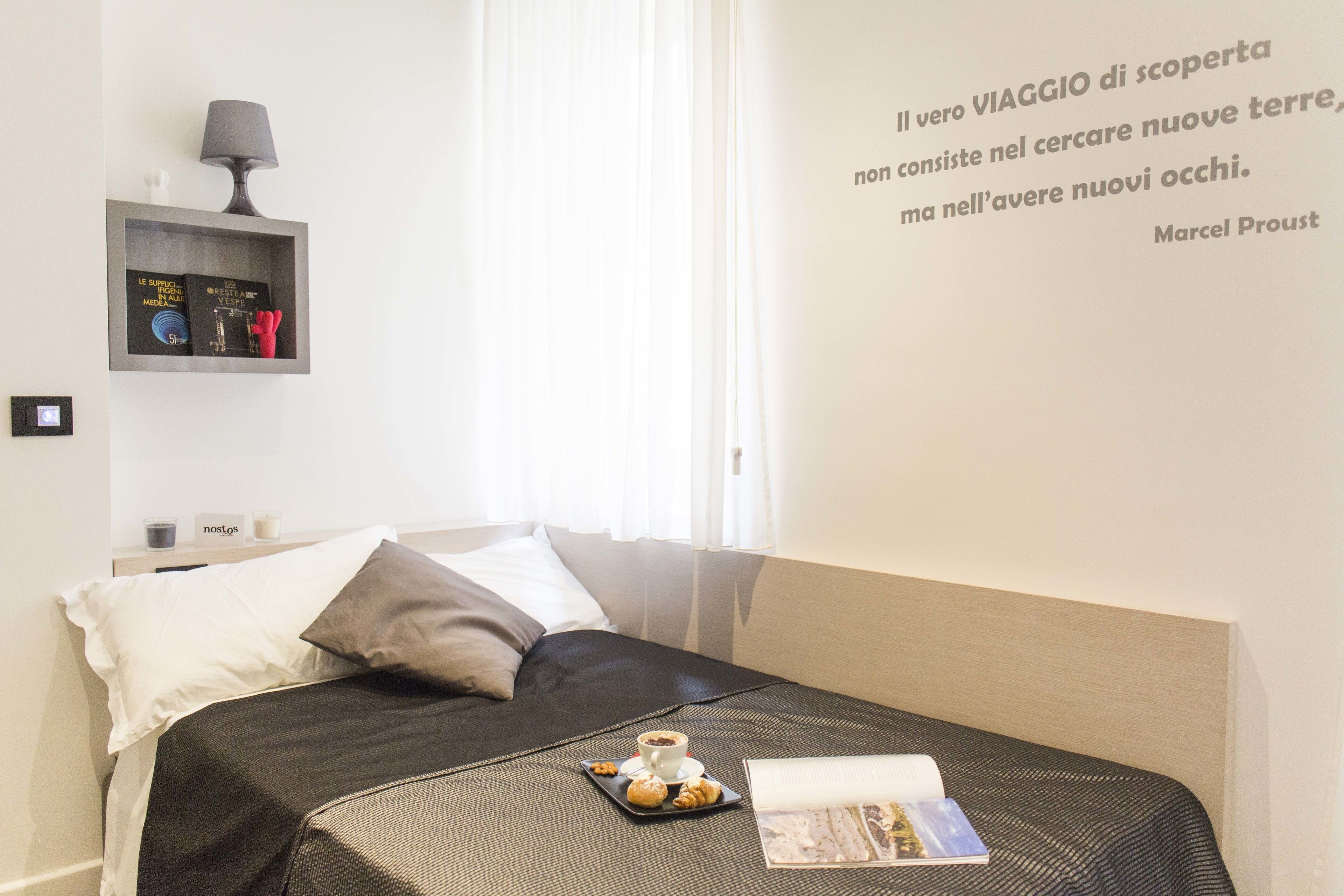 A neatly made bed with a tray of pastries and a book, a bedside lamp, and a shelf with books and decorative items.