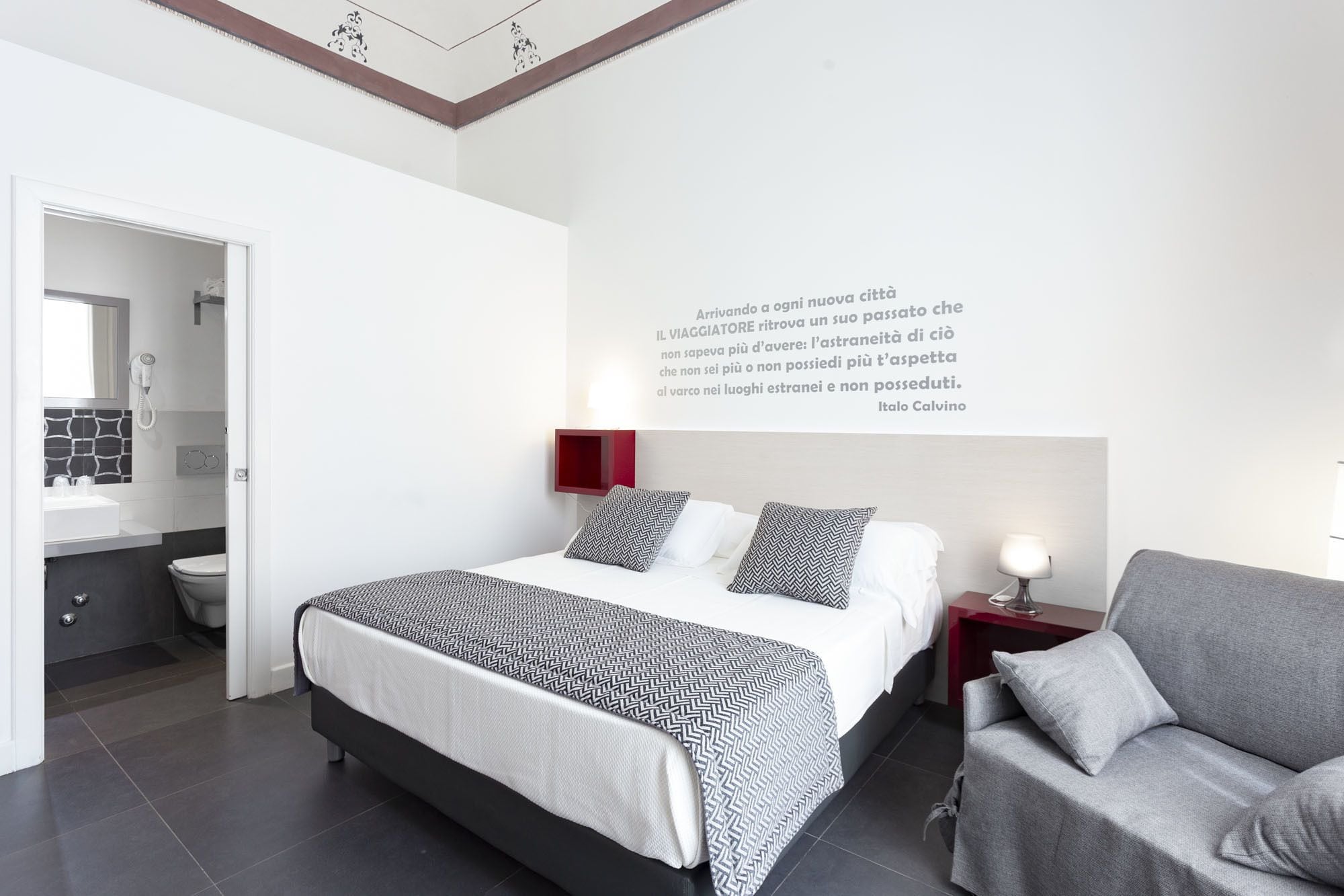 A modern bedroom with a bed, bedside tables, a chair, a wall-mounted lamp, and a bathroom visible through an open door.