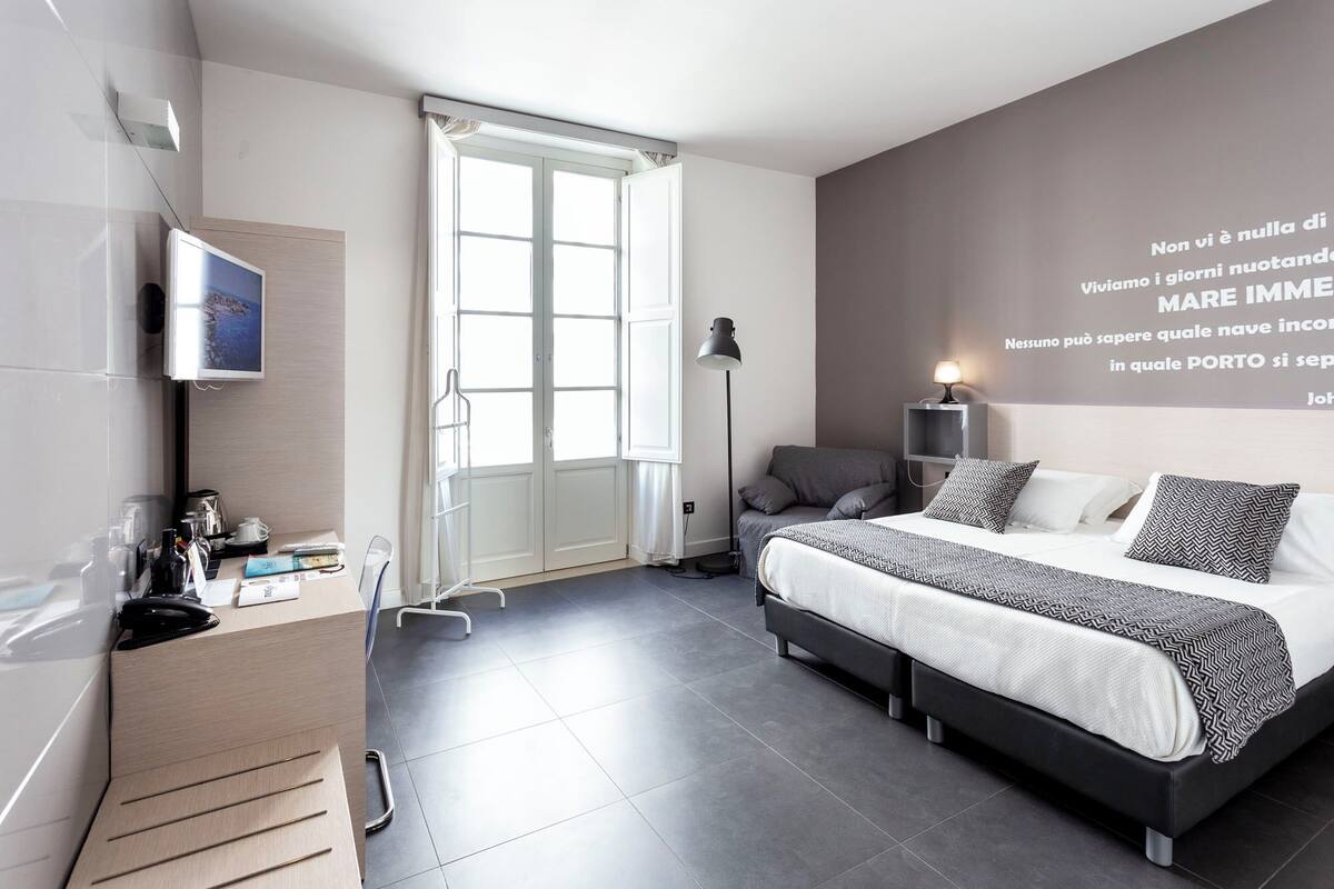 A modern bedroom with a large bed, a desk with a computer, a standing lamp, and a wall with a motivational quote.