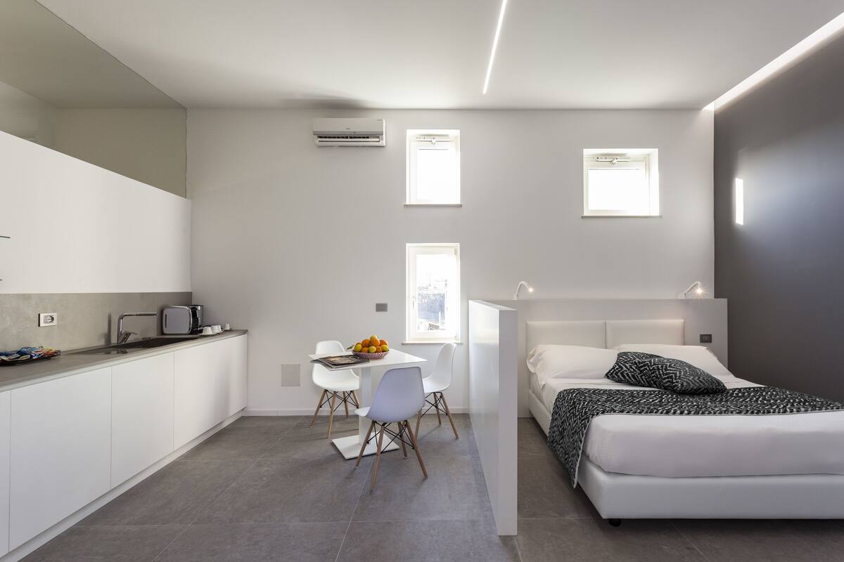 A modern, compact bedroom with a bed, bedside table, and a small desk with a chair. There is a window providing natural light, and the room has a neutral color palette.