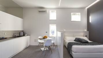 Un dormitorio moderno y funcional con cama, mesita de noche y un pequeño escritorio con silla. Una ventana aporta luz natural, y la habitación presenta una paleta de colores neutros.