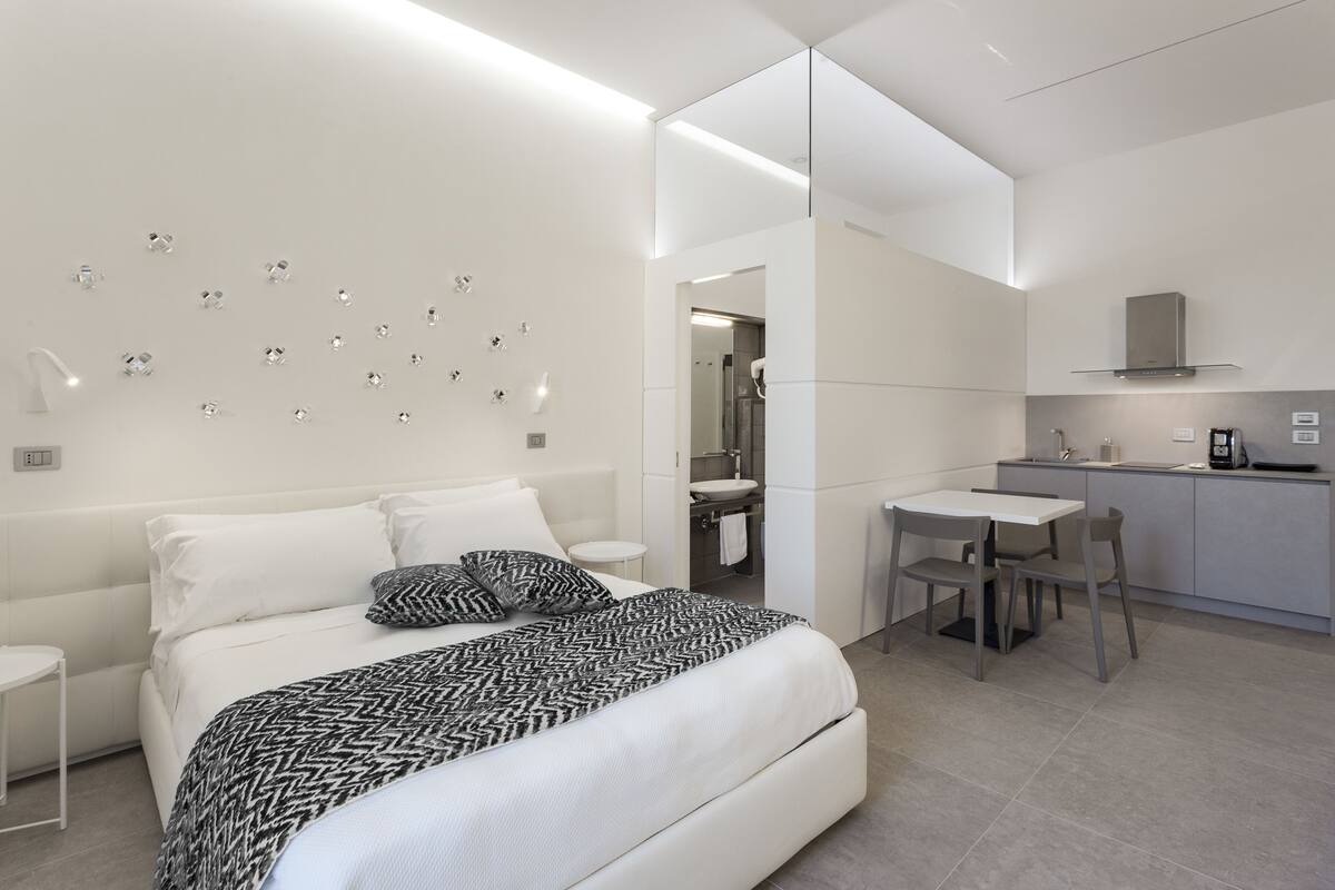 A modern hotel room with a bed, bedside table, and a small desk with a chair. There is a kitchenette with a sink and a stove.