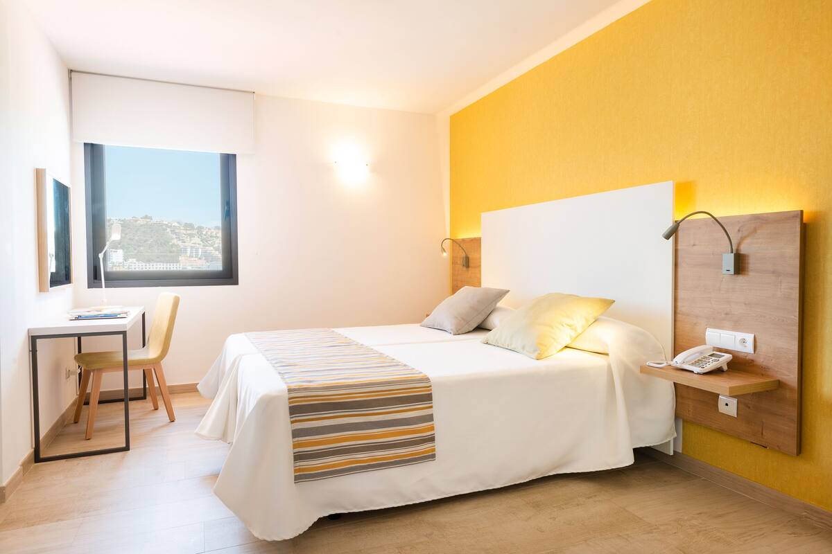 Een hotelkamer met een bed, een bureau, een stoel, een nachtkastje met telefoon en een raam met uitzicht op een stadsgezicht.