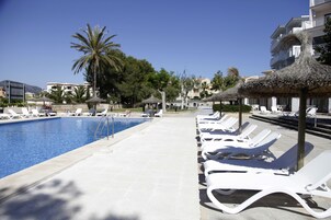 Zona de piscina con sillones de playa y sombrillas, rodeada de palmeras y edificios.