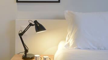 A neatly made bed with white linens, a bedside table with a lamp, two mugs, and a remote control.