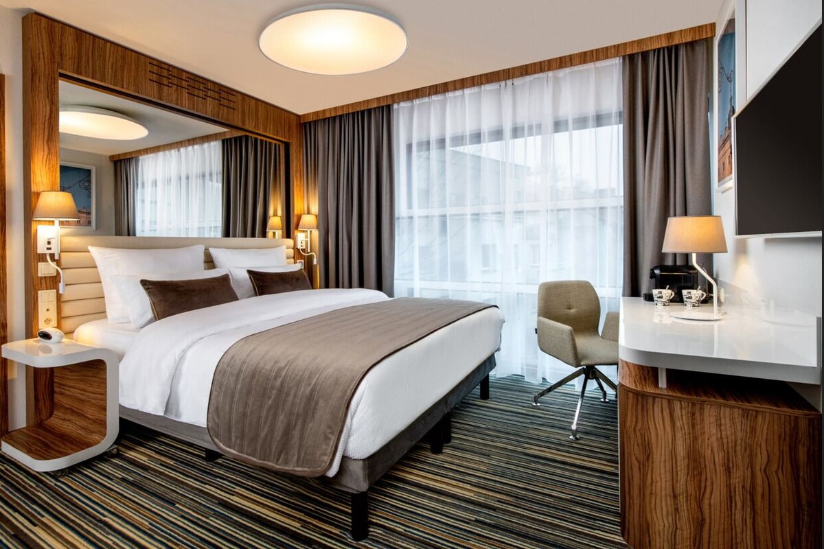 Ein Hotelzimmer mit einem großen Bett, einem Schreibtisch, einem Sessel, einem Spiegel und einem fenster mit Vorhängen.