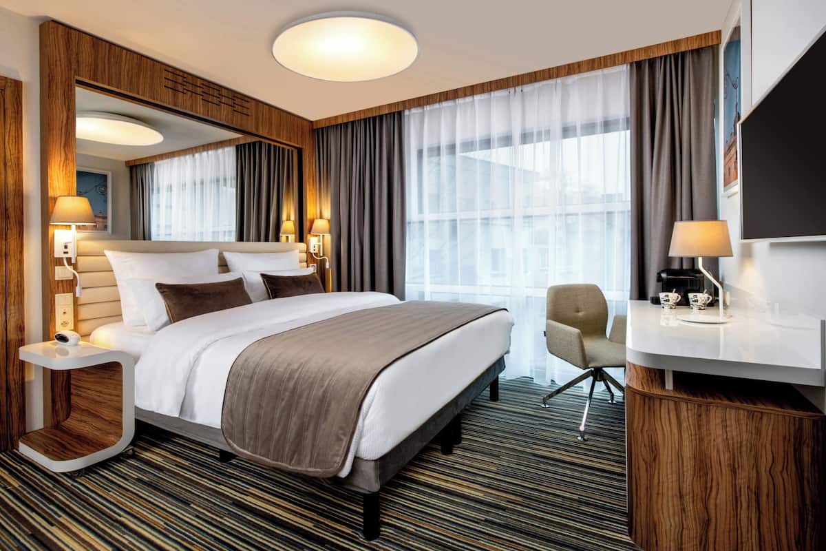 Hotellihuone, jossa on suuri sänky, yöpöydät lamppuineen, työpöytä tuolilla sekä suuri ikkuna, jossa on verhot.