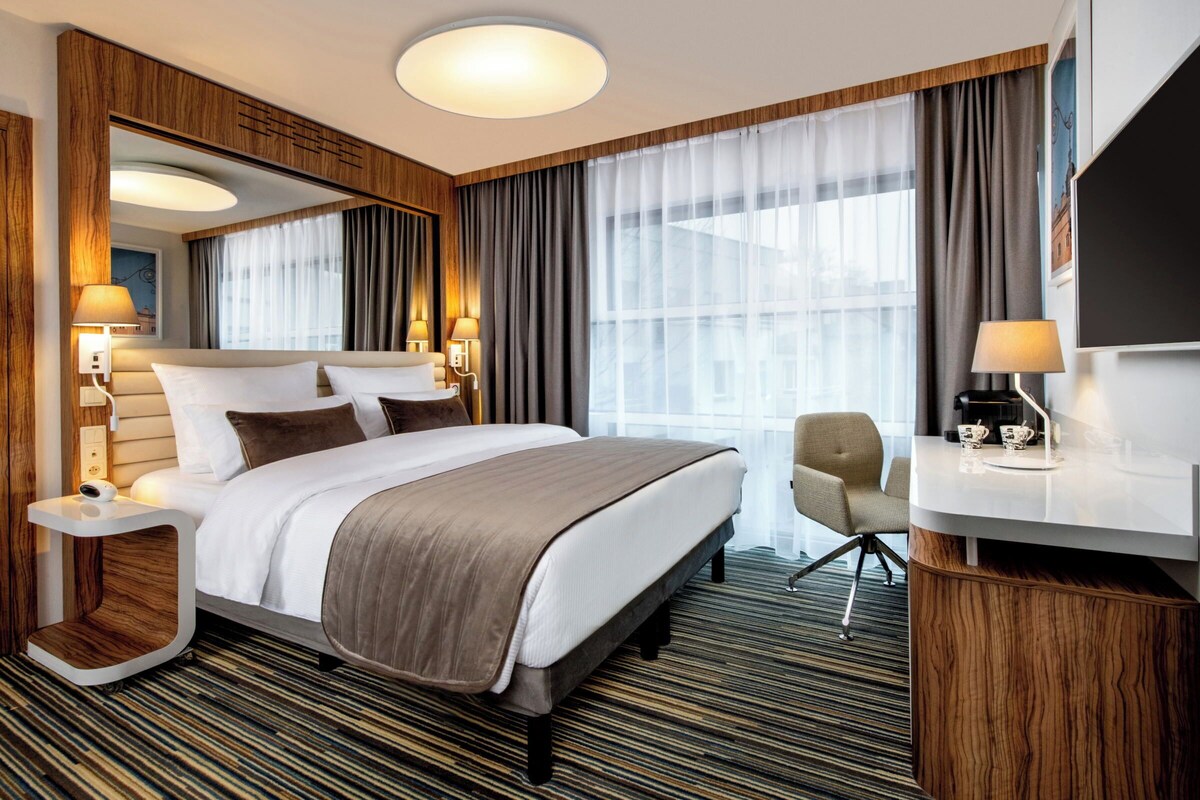 Ein Hotelzimmer mit einem großen Bett, Nachttischen, einem Schreibtisch, einem Sessel und einem großen Fenster mit Vorhängen.