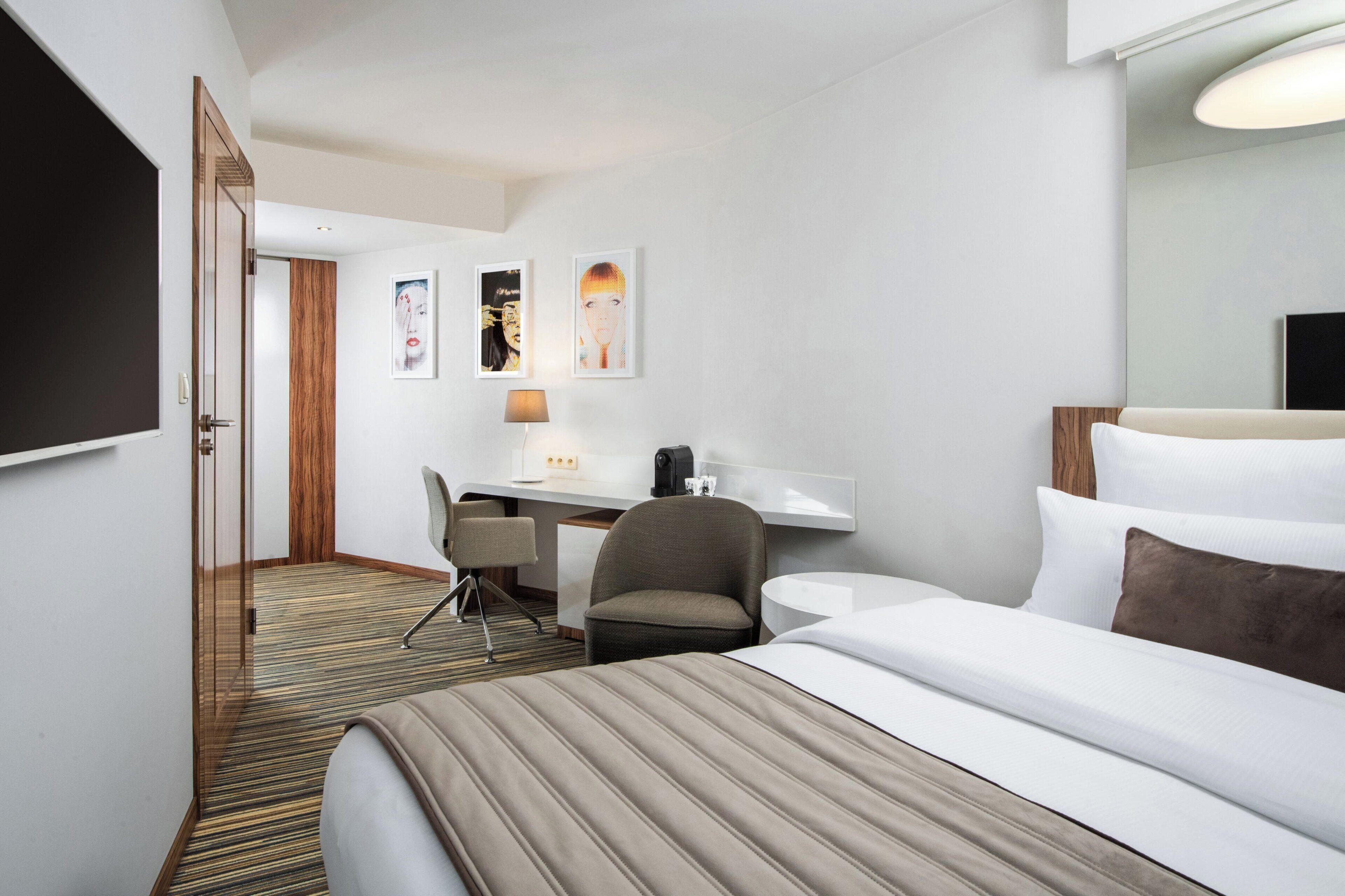 A hotel room with a bed, a desk, a chair, a desk lamp, and a flat-screen TV.
