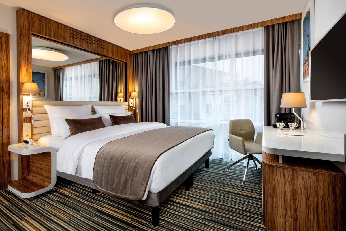 A hotel room with a large bed, bedside tables with lamps, a desk with a chair, and a large window with curtains.