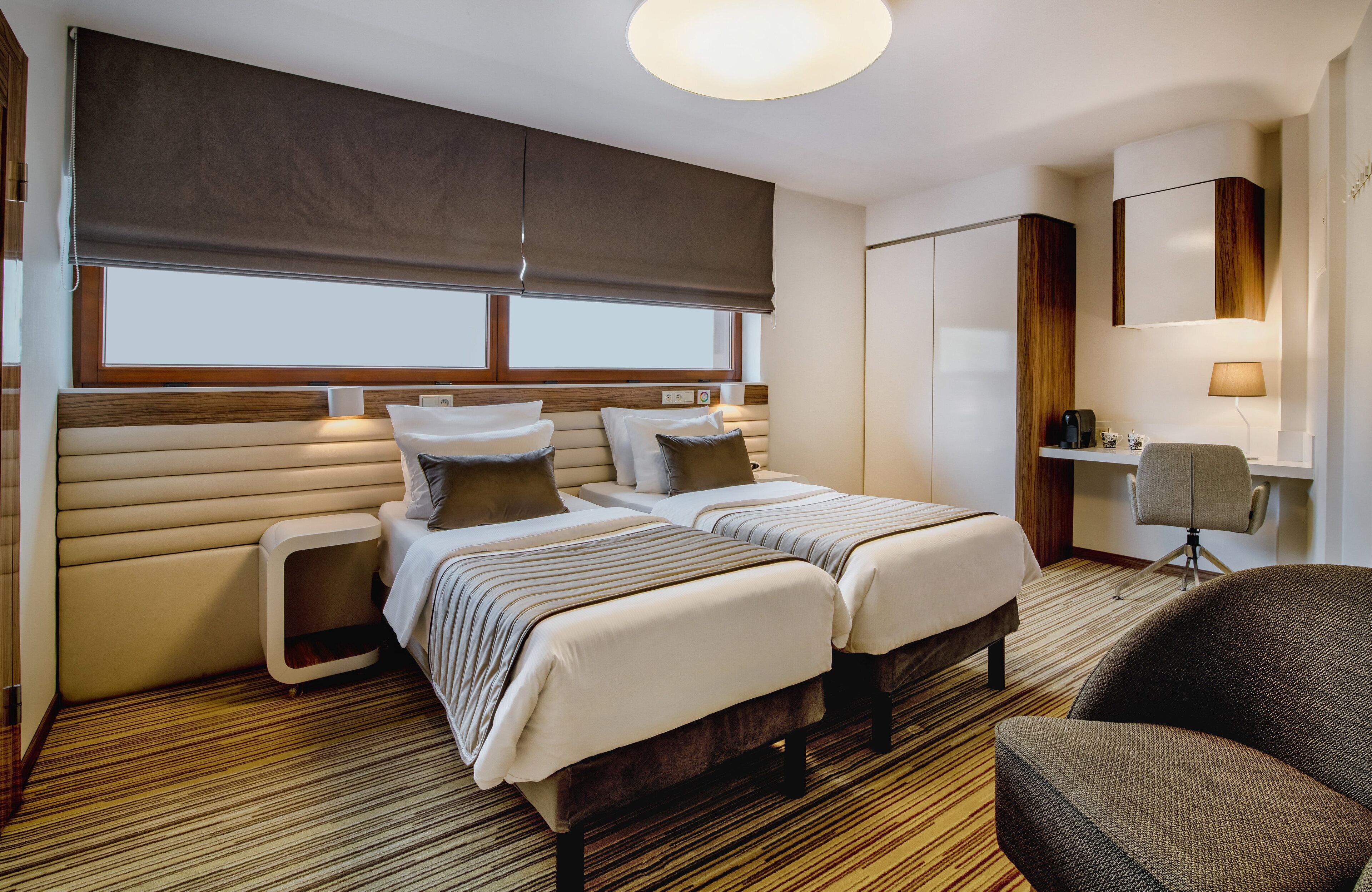 A hotel room with two beds, a desk, a chair, and a large window with blinds.